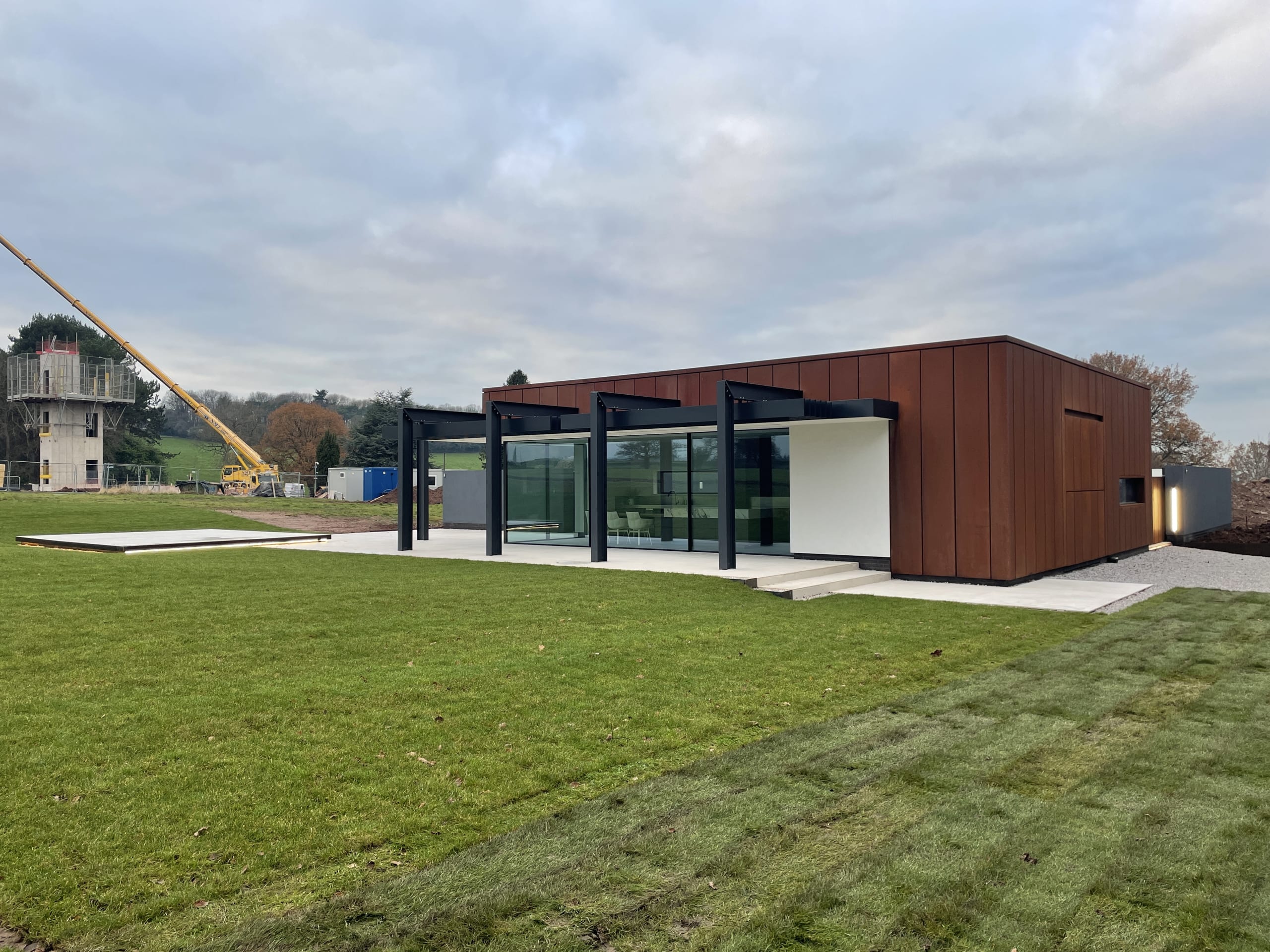 Modern single-story building with Corten steel cladding, black steel pergola, and large glass walls set in green lawn